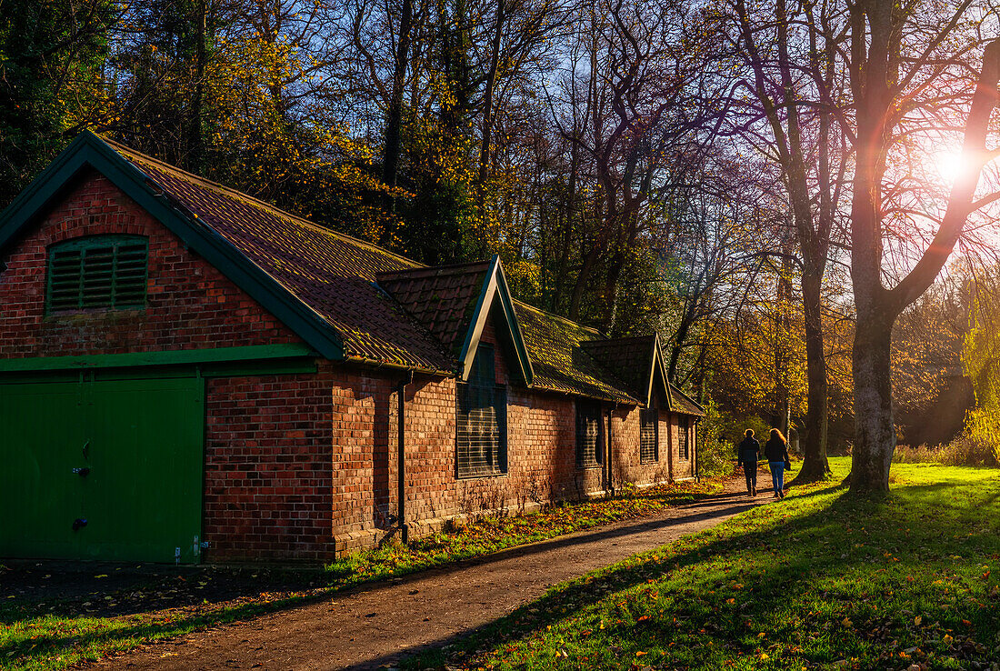 Freunde spazieren auf einem Weg vorbei an einem Backsteingebäude in einem Park in Durham bei Sonnenuntergang,Durham,Durham,England