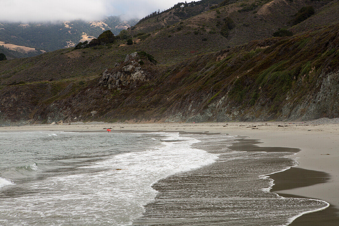 Waves and surf come ashore on the Big Sur coast. A lone swimmer,boarder,stands in the water.,Big Sur,California