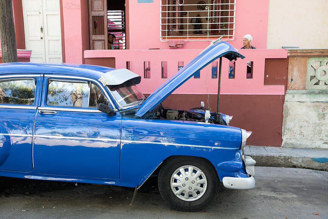 Auf einer Straße in der Innenstadt von Havanna wird die Motorhaube eines amerikanischen Oldtimers zur Wartung angehoben, während die Leute um ihr Haus herum sitzen,Havanna,Kuba