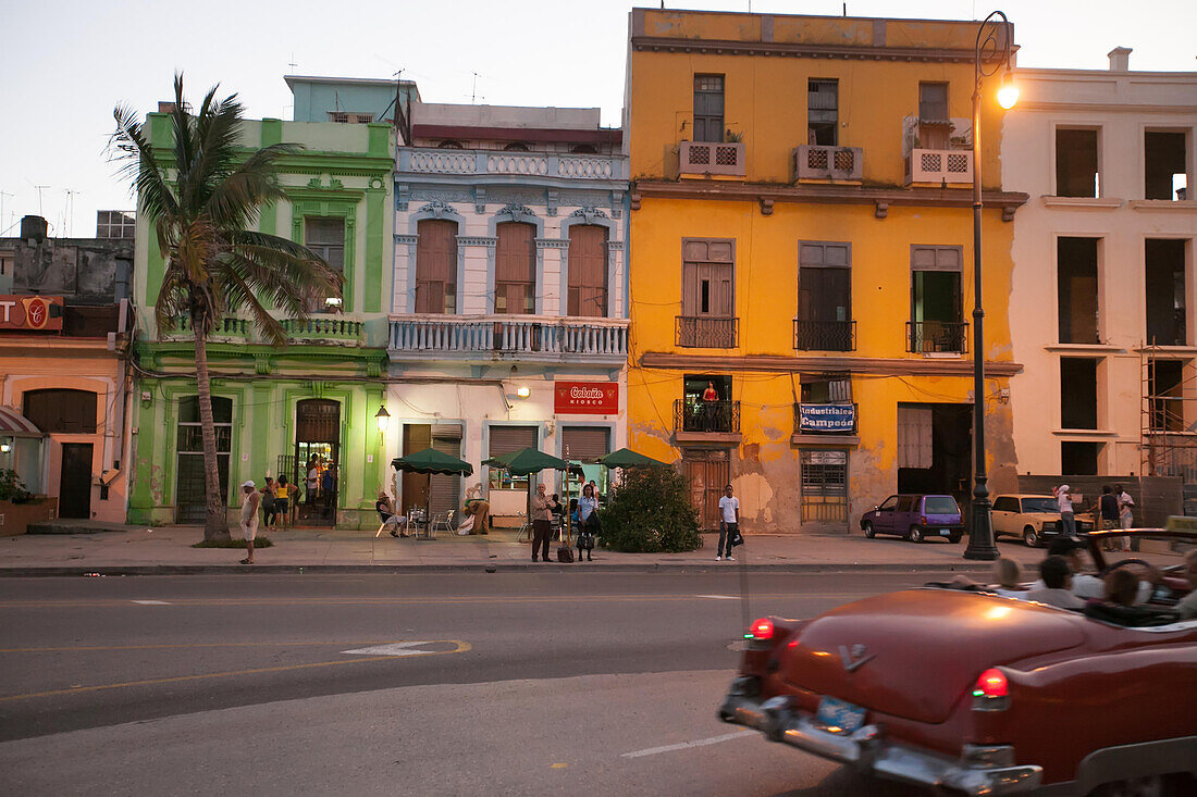 In der Abenddämmerung stehen Menschen auf der Straße, als ein amerikanischer Oldtimer in der Innenstadt von Havanna vorbeifährt.,Havanna,Kuba