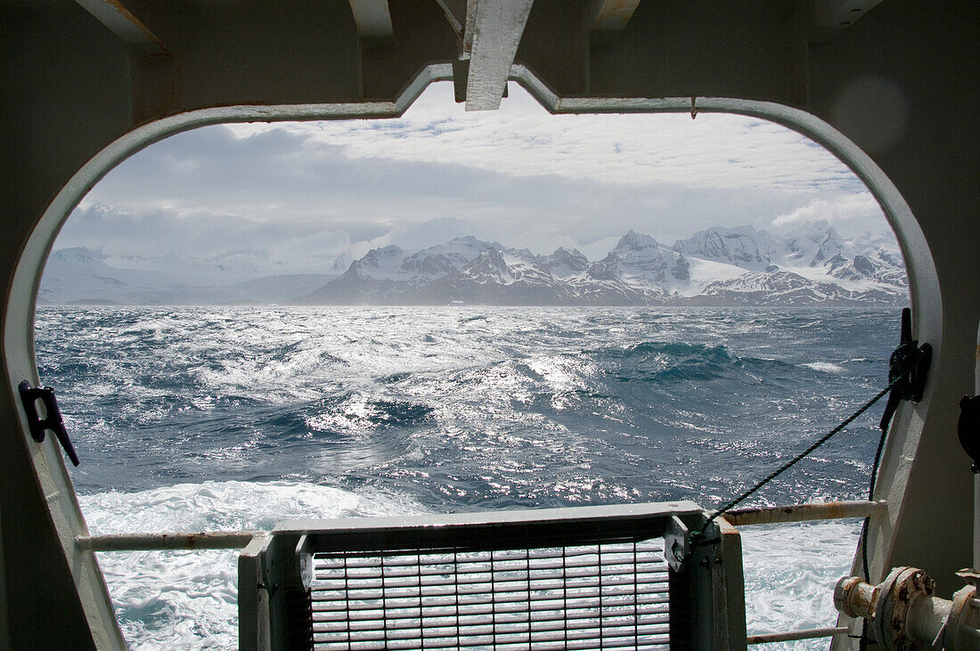 Blick aus dem Fenster eines Schiffes auf die Wellen des Südlichen Ozeans und die schneebedeckten Berge der Insel Südgeorgien an einem sonnigen Tag, Südgeorgien, Antarktis