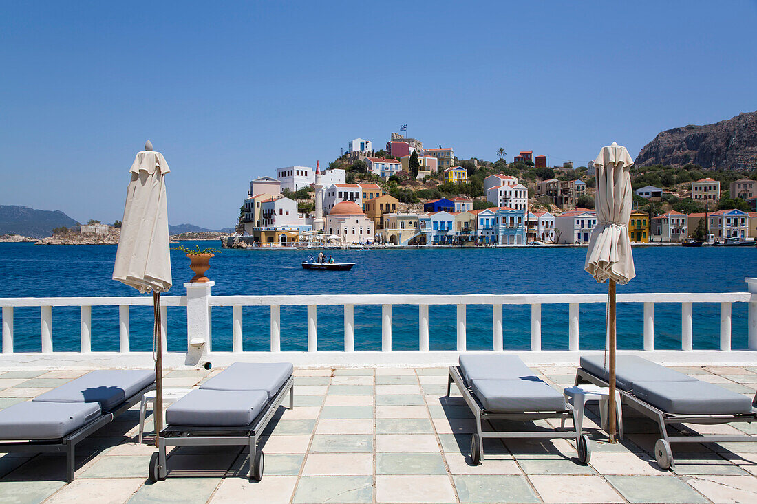 Liegestühle und Sonnenschirme auf einer Hotelterrasse mit Blick auf den Hafen und die farbenfrohen Gebäude entlang der Küstenlinie auf der anderen Seite des Hafens, Kastellorizo, Dodekanes, Griechenland