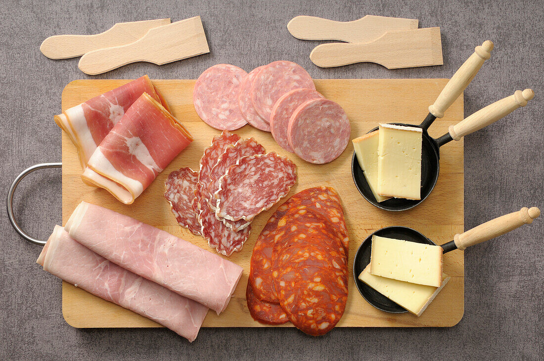 Overhead-Ansicht von Fleisch und Käse für Raclette auf Schneidebrett auf grauem Hintergrund, Studio Shot