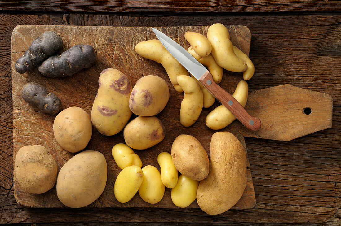 Draufsicht auf verschiedene Kartoffelsorten auf Schneidebrett mit Messer