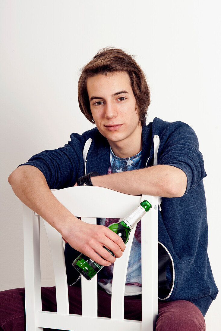 Porträt eines Teenagers, der auf einem Stuhl sitzt, eine Bierflasche in der Hand hält, lächelt und in die Kamera schaut, Studioaufnahme auf weißem Hintergrund