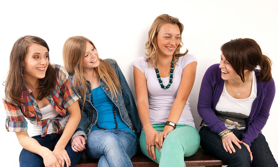 Four,young women sitting on bench together,laughing and looking at each other,studio shot on white background