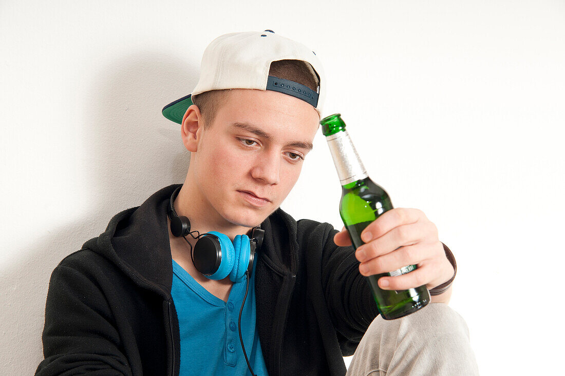 Close-up of teenage boy,sitting and leaning against wall,wearing hat and headphones around neck,holding bottle of beer,studio shot on white background