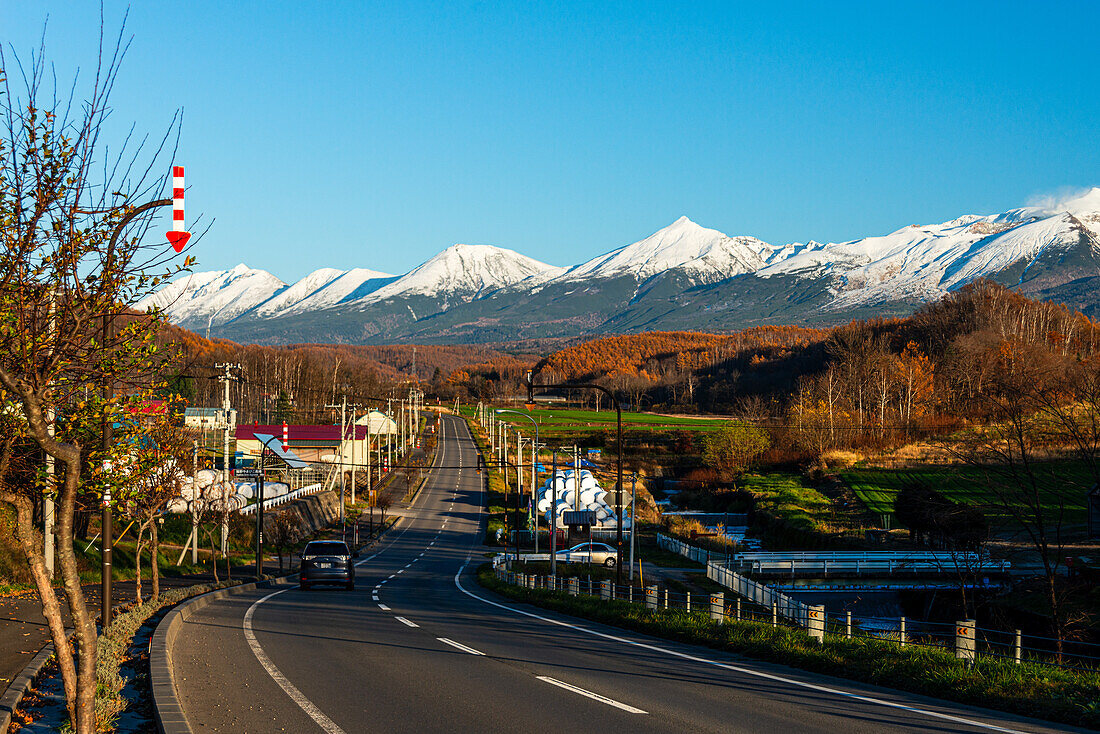 Alpenpanorama einer ländlichen Gegend mit einer Straße, die zum verschneiten Tokachi-Gebirge in Hokkaido, Japan, Asien führt