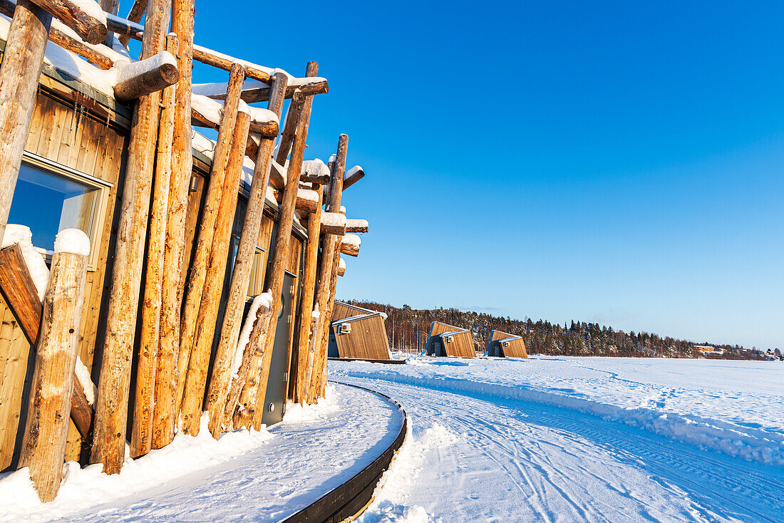Architektonische Struktur des arktischen Badehotels aus Baumstämmen auf dem gefrorenen Fluss Lule mit hochmodernen Chalets im Hintergrund, Harads, Norrbotten, Schwedisch-Lappland, Schweden, Skandinavien, Europa