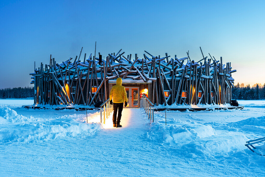 Person steht auf der Brücke, die das Hauptgebäude des beleuchteten Arctic Bath Hotels aus Holzstämmen verbindet, in der Abenddämmerung, Harads, Schwedisch-Lappland, Norrbotten, Schweden, Skandinavien, Europa