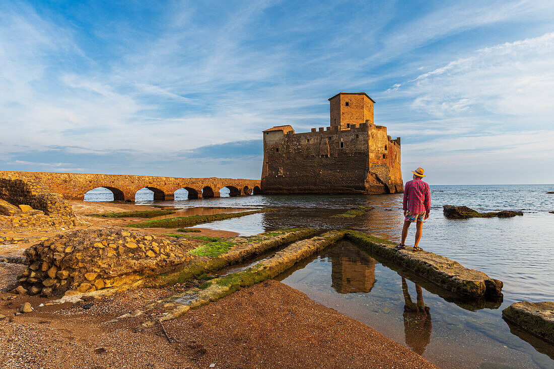 Mann mit Hut steht auf einer römischen Ruine mit Blick auf die mittelalterliche Burg von Torre Astura, die aus dem Meer ragt, Gemeinde Nettuno, Provinz Rom, Tyrrhenisches Meer, Latium (Lazio), Italien, Europa