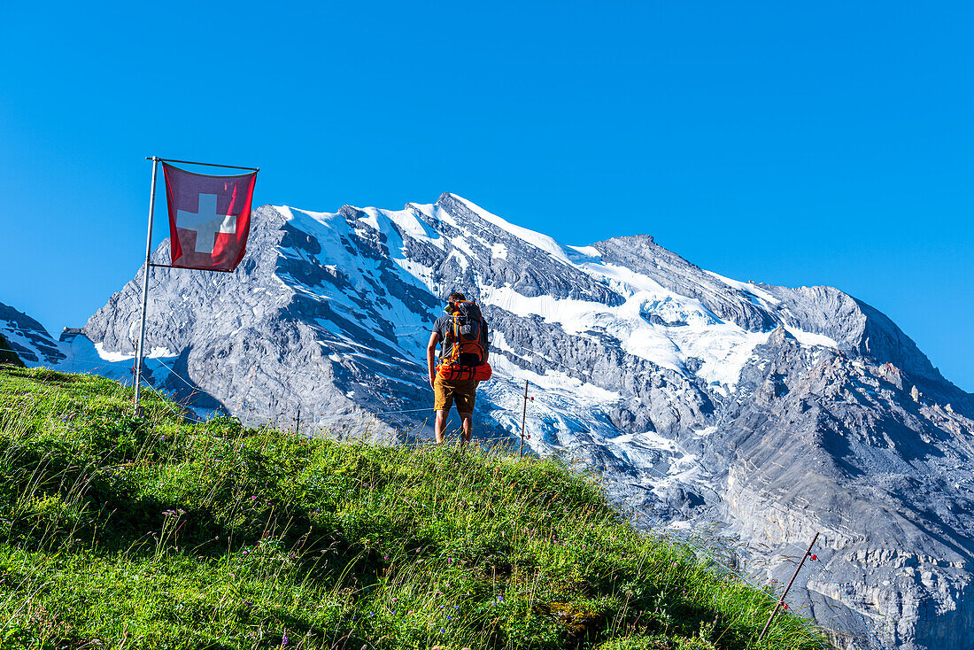 Rear view of a man with backpack hiking the Swiss Alps passing a Swiss flag flying,Oeschinensee,Kandersteg,Bern Canton,Switzerland,Europe