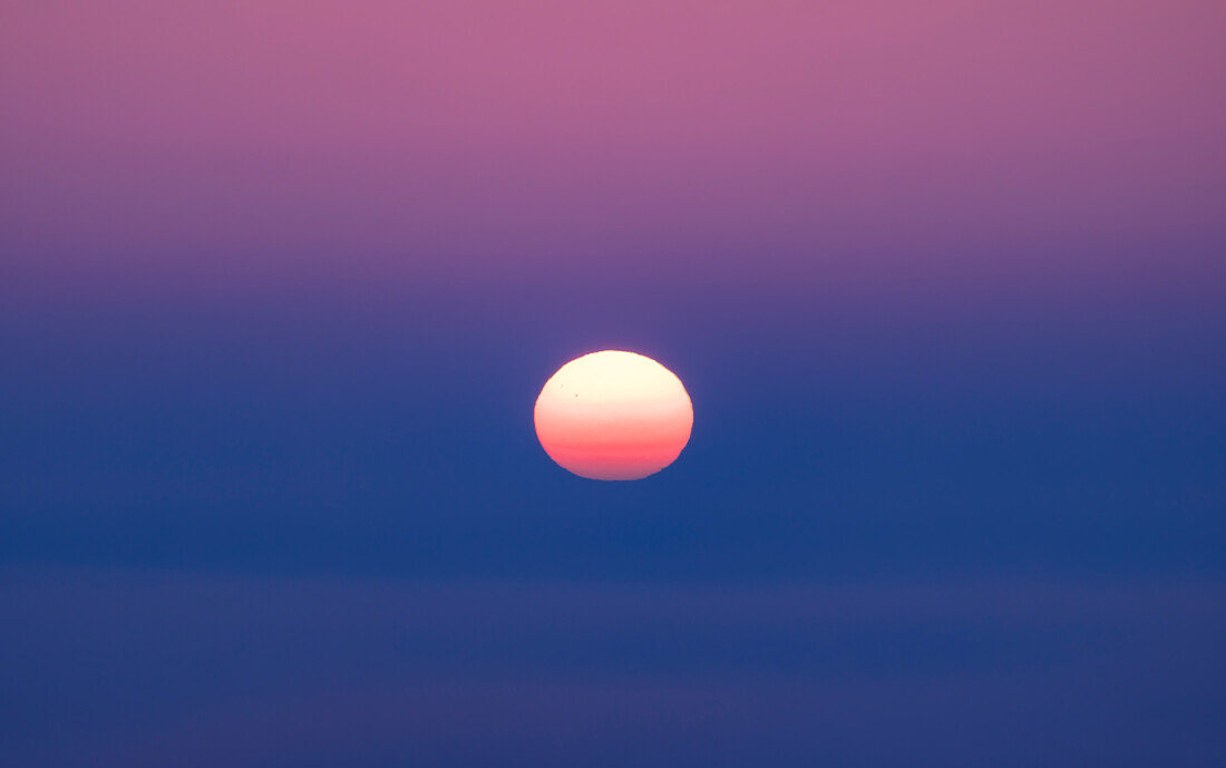 Die untergehende Sonne taucht in eine Nebelbank ein und blickt nach Westen, am Abend der Frühlings-Tagundnachtgleiche, 20. März 2023. Der Nebel verdunkelt und rötet die Sonne, was die atmosphärische Absorption und Brechung zeigt, da die Sonnenscheibe recht flach ist. Ein paar Sonnenflecken sind gerade noch sichtbar. Der Farbverlauf ist natürlich und ergibt sich aus der Mischung von Nebel unten und klarerem Dämmerungshimmel oben.