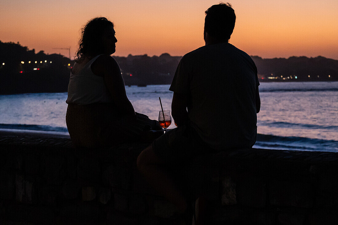 Ein Paar genießt einen Drink bei Sonnenuntergang an der Promenade Jacques Thibaud vor dem Strand Grande Plage in Saint Jean de Luz, einem Fischerdorf an der Mündung des Flusses Nivelle im südwestfranzösischen Baskenland.