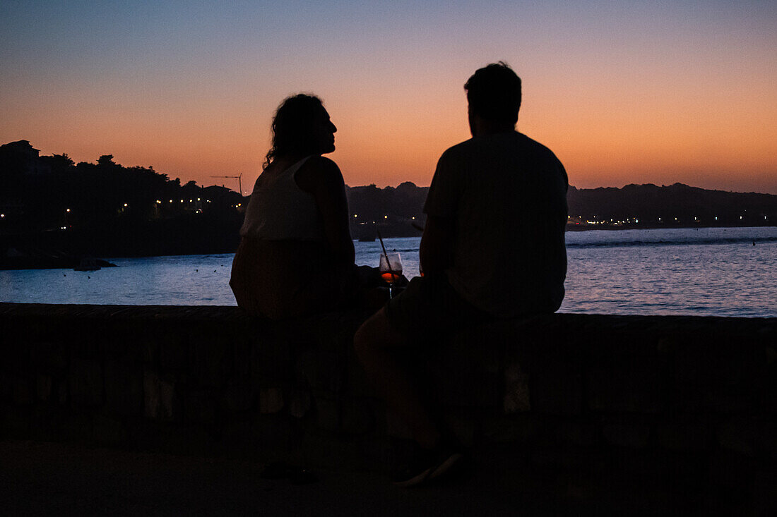 Ein Paar genießt einen Drink bei Sonnenuntergang an der Promenade Jacques Thibaud vor dem Strand Grande Plage in Saint Jean de Luz, einem Fischerdorf an der Mündung des Flusses Nivelle im südwestfranzösischen Baskenland