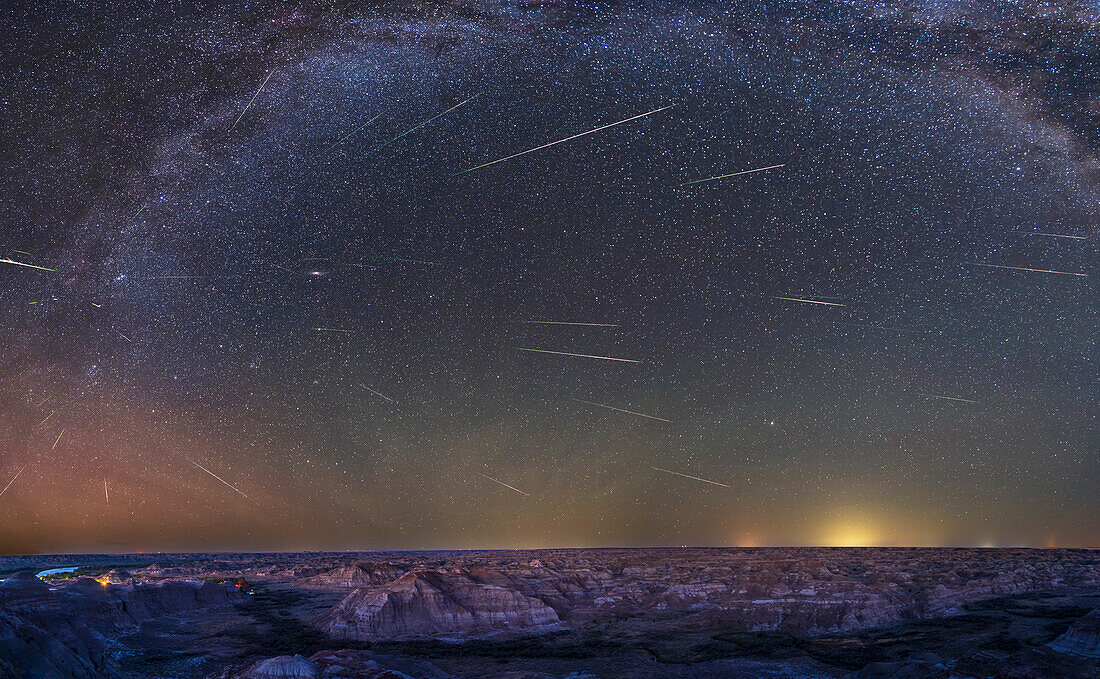 This is a panorama of the ground and sky framing the badlands of Dinosaur Provincial Park,in the Red Deer River valley in Alberta,with the summer sky above filled with three dozen meteors from the annual Perseid meteor shower,of August 12,2023. The meteors all appear to be streaking away from the radiant point in Perseus at far left. The location is the upper viewpoint near the Park Entrance,and looking northeast to southeast. The lights in the valley below are from the campground and service centre. The one glaring sodium vapour light should be removed or at least shielded. The brightest hori