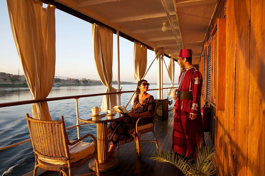 Egypt, Upper Egypt, Nile Valley, tourist sitting in a passageway of Steam Ship Sudan, the last steamboat cruising on the Nile, alongside a crew member in period costume and wearing a hat. tarbouche