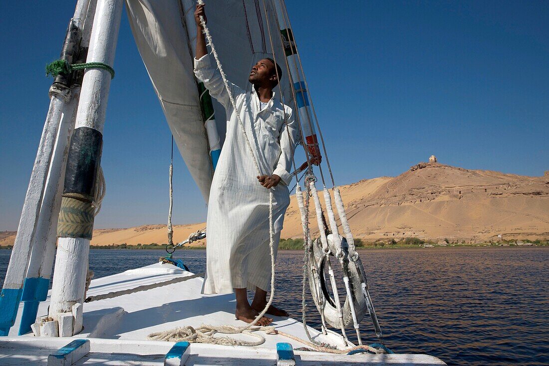 Egypt, Upper Egypt, Aswan, Nubian sailor in front of his felucca all sails erected sailing on the Nile in front of a high sand dune of the left bank