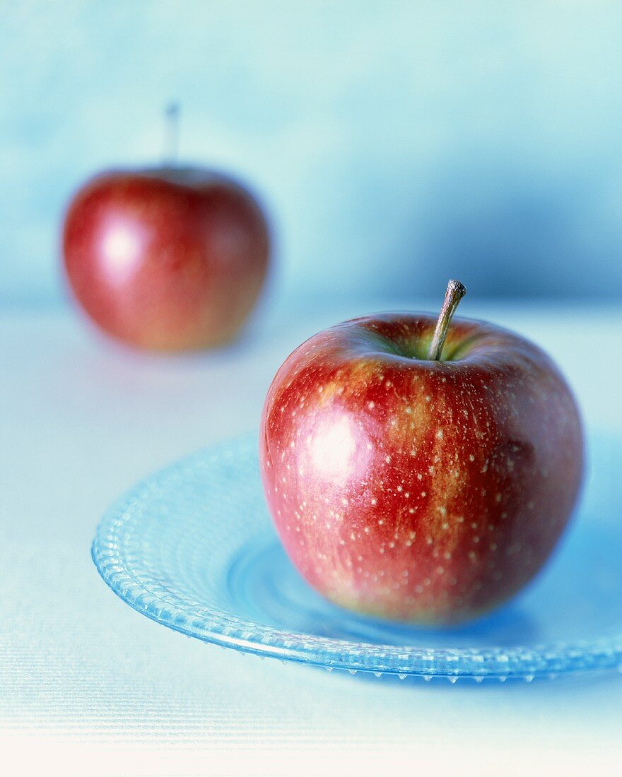 Zwei rote Äpfel, einer im Vordergrund auf blauem Teller
