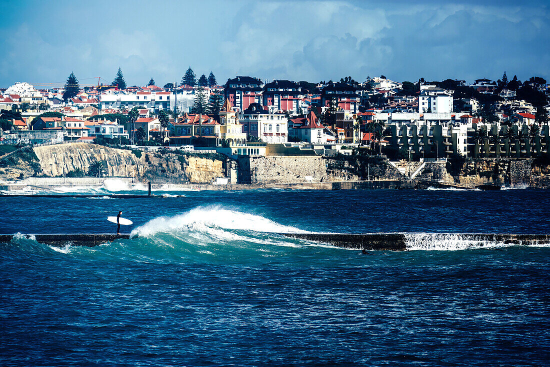 Surfer auf einem Pier bei stürmischer See mit Cascais-Estoril-Gebäuden im Hintergrund kurz vor Lissabon,Portugal,Europa