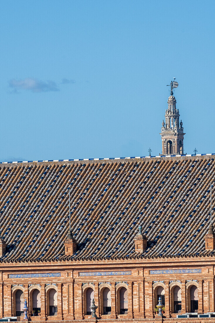 A vibrant skyline view showing La Giralda tower rising behind a historic rooftop in Seville, Spain. Bright blue sky complements the architectural details of Plaza de Espana.