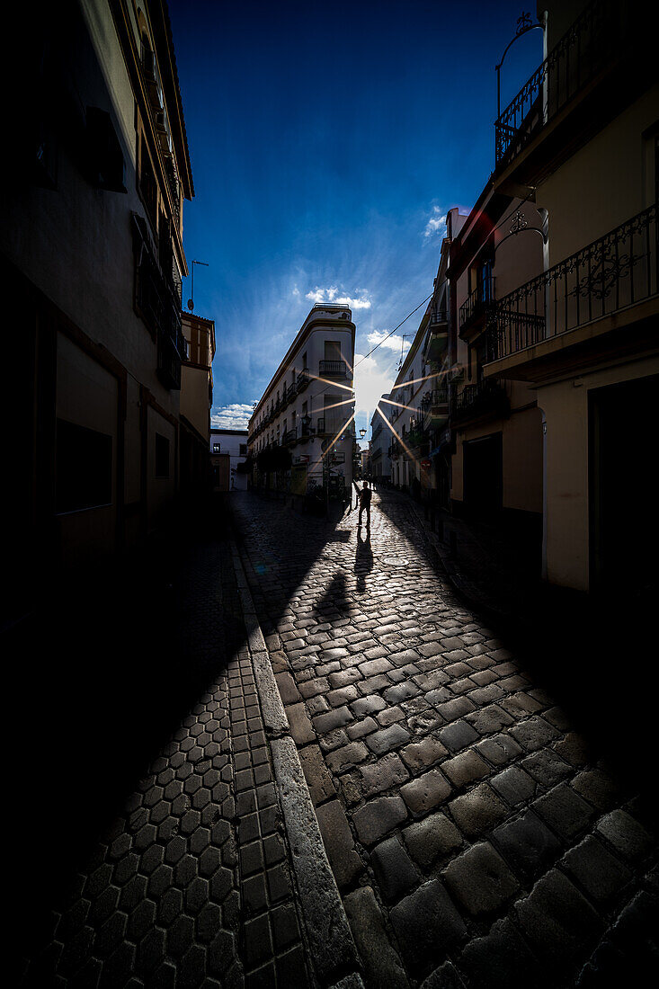 A beautiful sunset casts dramatic shadows in the historic center of Sevilla, Spain. The sun gently illuminates the cobblestone street of Calle Feria, creating a stunning urban atmosphere.