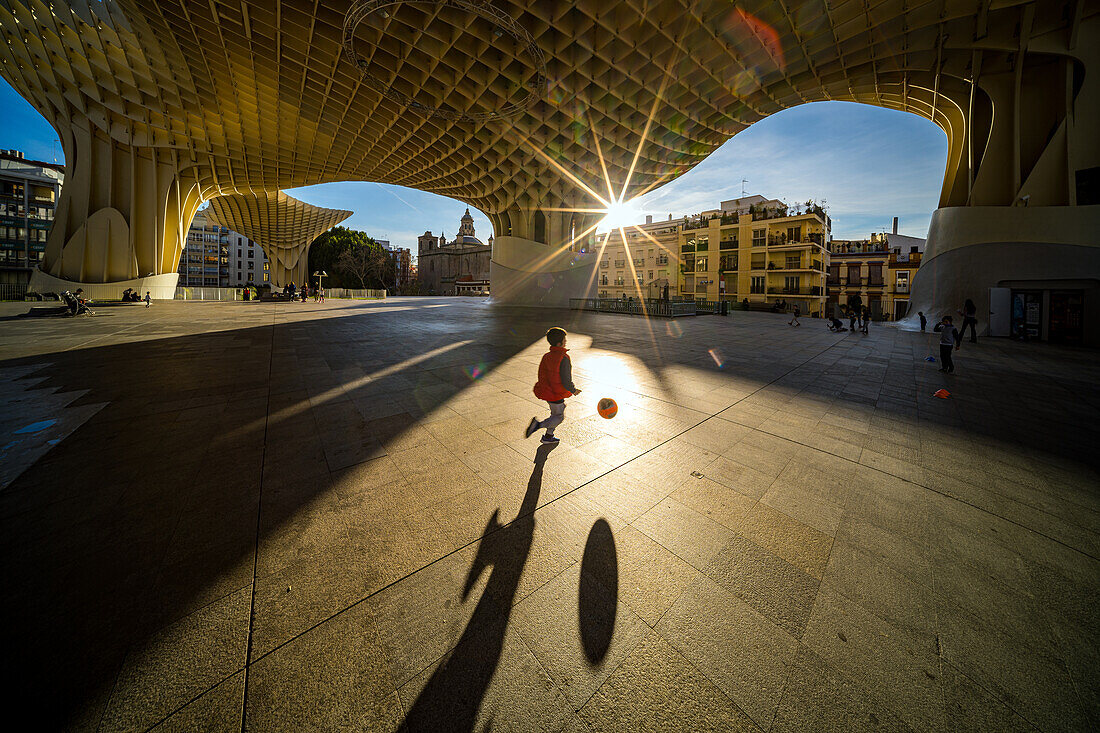 Ein kleines Kind spielt vergnügt mit einem Ball unter dem ikonischen Bauwerk Las Setas in Sevilla,Spanien. Die architektonische Schönheit und die sonnenbeschienene Umgebung schaffen ein lebendiges Stadtbild.