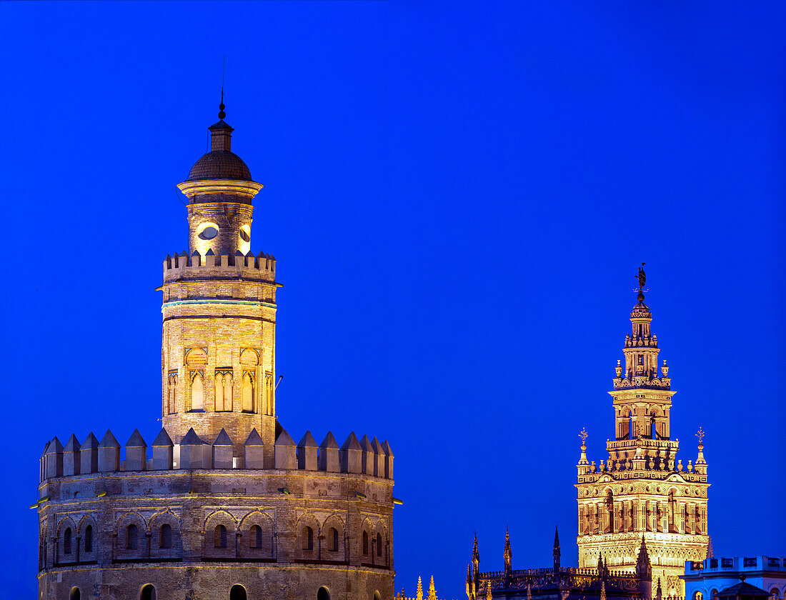 A nighttime view of Seville featuring the Torre del Oro and La Giralda under a vibrant blue sky. The photograph captures the historic beauty and architectural splendor of these iconic landmarks.