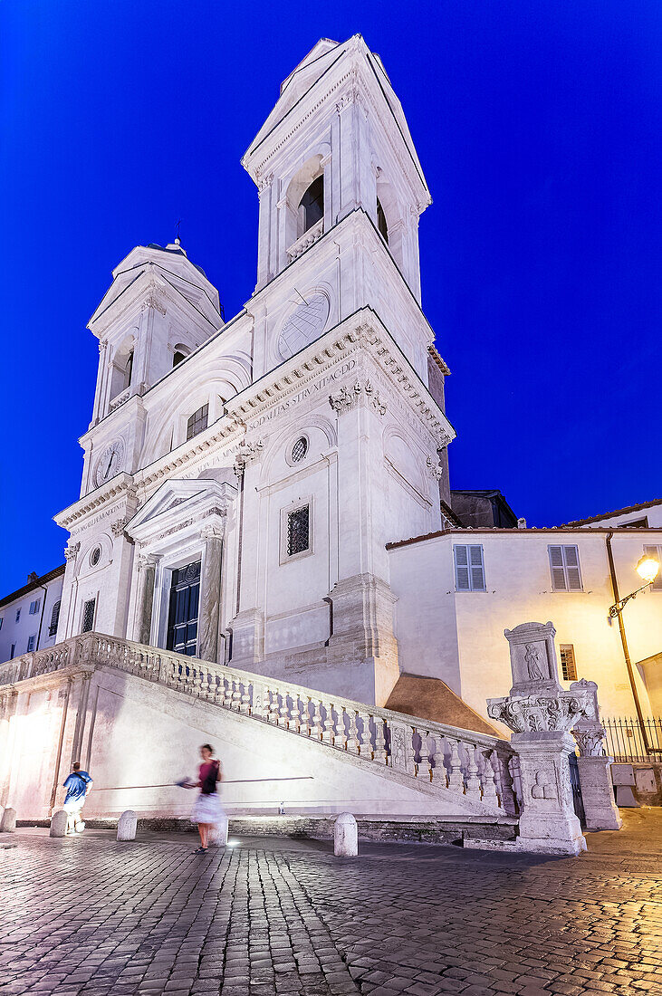 People stroll by the stunning Trinità dei Monti, illuminated against the night sky in Rome, creating a charming evening atmosphere.