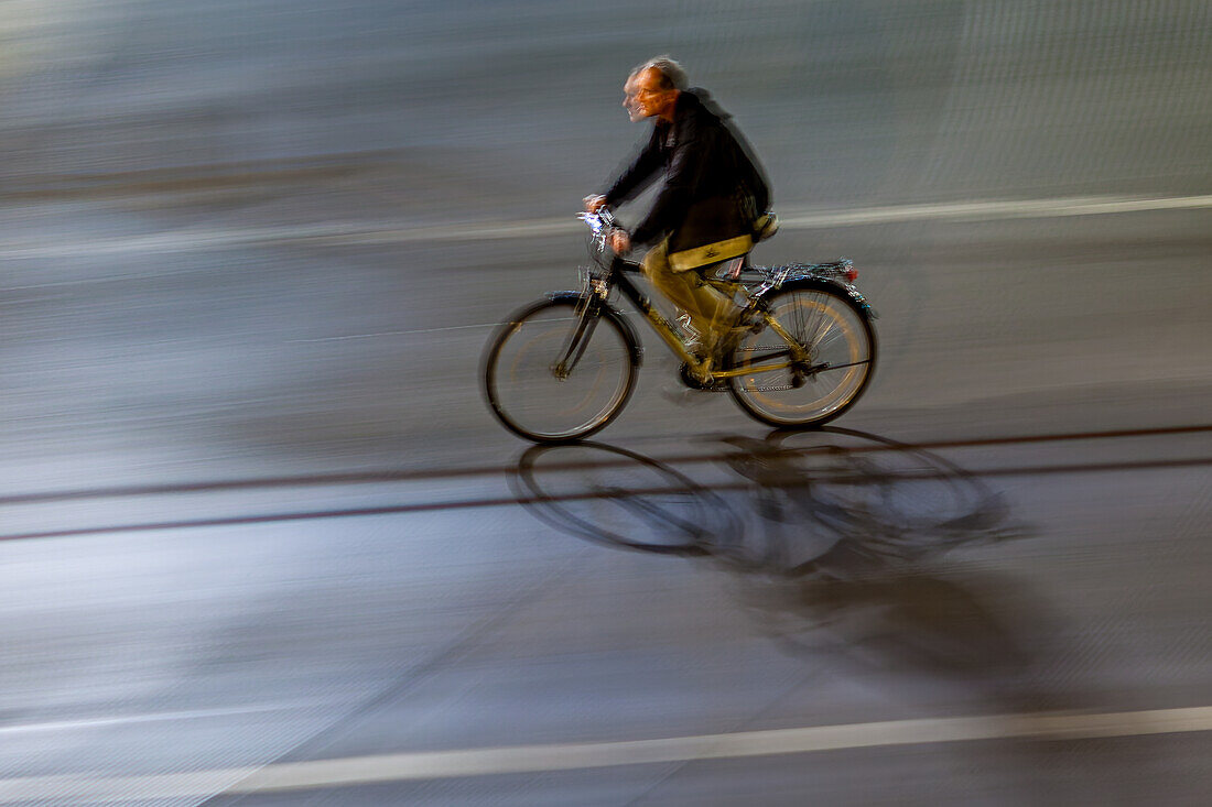 A man riding a bicycle on city streets at night, captured with a motion blur effect for dynamic movement.