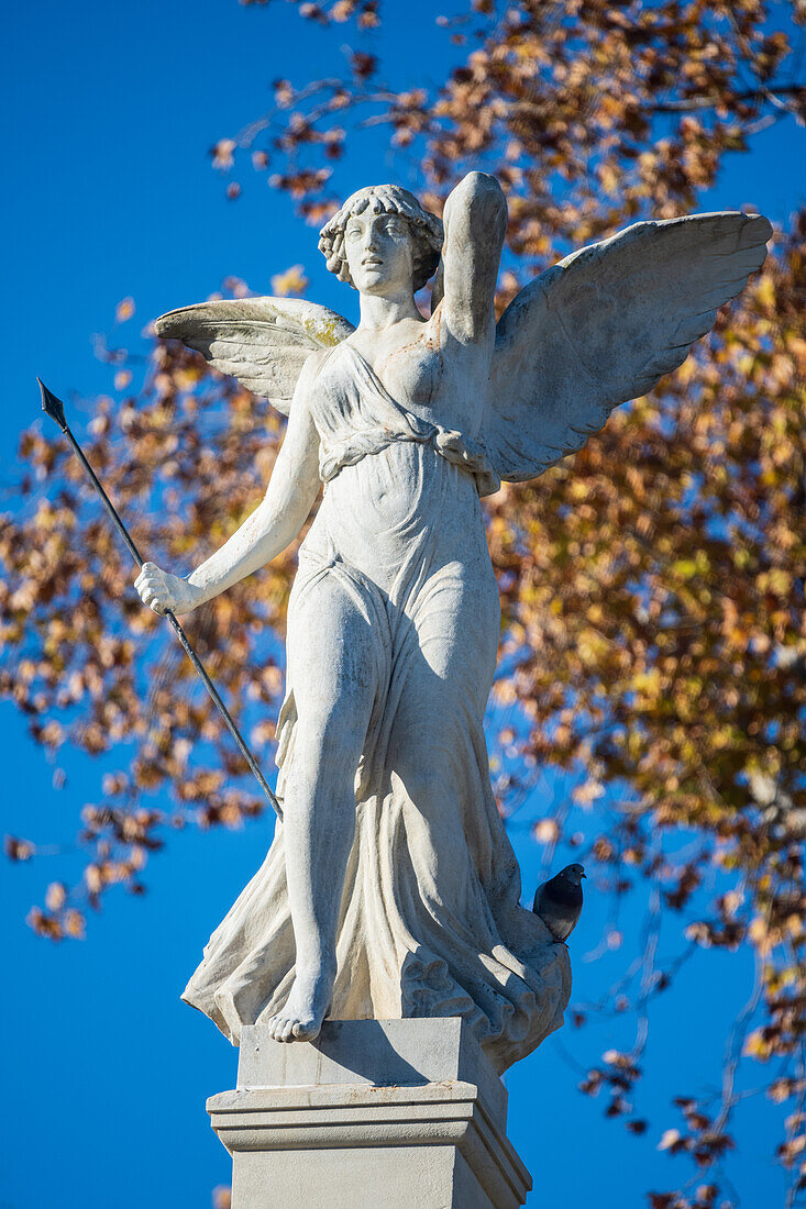 Die Victoria-Alada-Statue auf der Plaza de America,Parque de Maria Luisa,Sevilla,Spanien. Das Bild zeigt das kulturelle Erbe und die Schönheit der Skulptur vor dem Hintergrund des bunten Herbstlaubs und des klaren blauen Himmels.