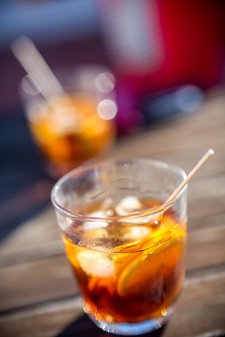 Eine Nahaufnahme von zwei Gläsern mit erfrischenden Wermut-Cocktails auf einem sonnenbeschienenen Terrassentisch,die die leuchtenden Farben und das einladende Ambiente hervorheben.