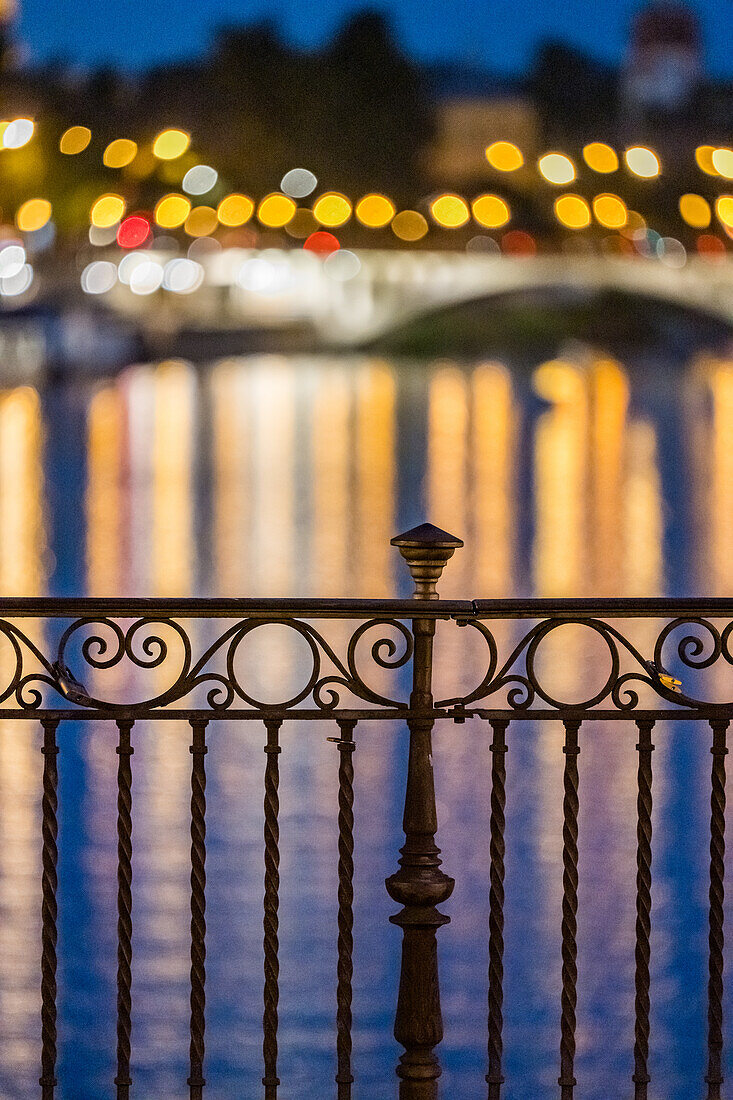 Intricate ironwork balustrade of Triana Bridge with colorful light reflections in a serene nighttime setting.