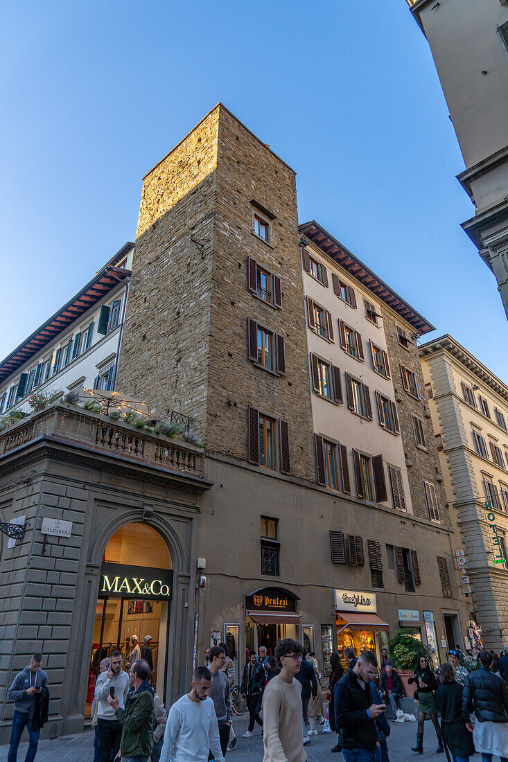 Ein mittelalterlicher Turm und moderne Geschäfte in einer Straße mit Touristen in Florenz, Italien.