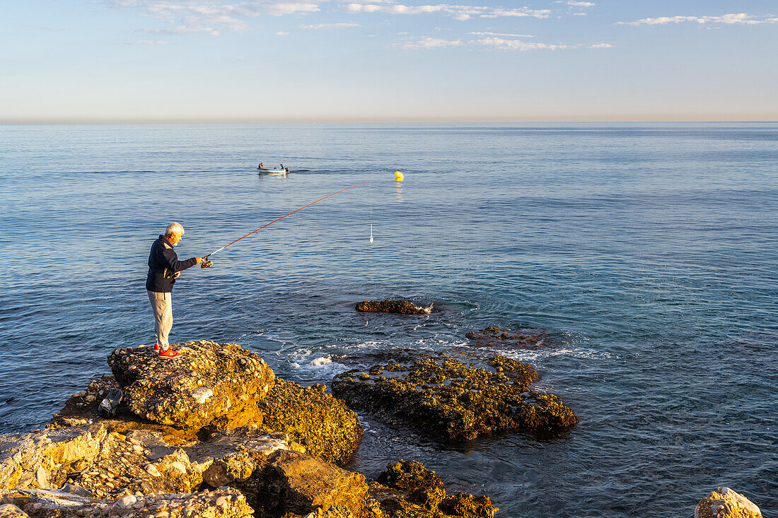 Nerja, Spain, May 5 2022, A man fishing from rocks near the sea with a nearby boat in Spain.