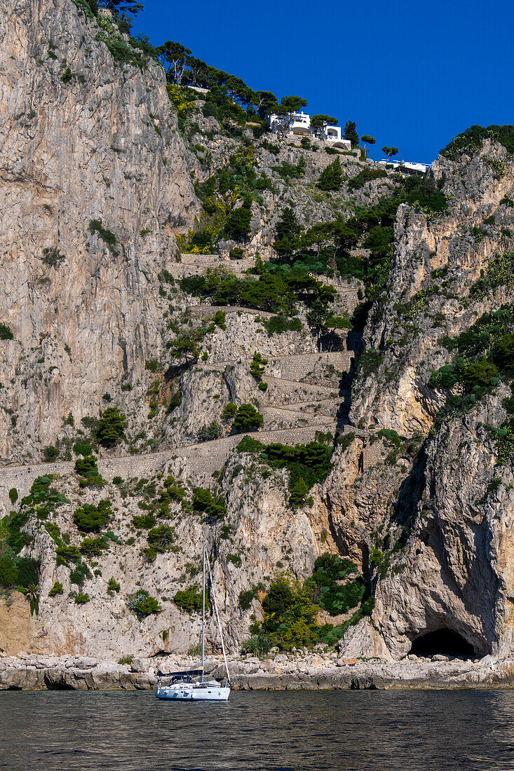 Ein Segelboot ankert in den Gewässern des Tyrrhenischen Meeres vor der Küste der Insel Capri, Italien. Oben sind die Villen der Stadt Capri zu sehen, während sich die Via Krupp in Serpentinen die Klippe hinunterschlängelt.