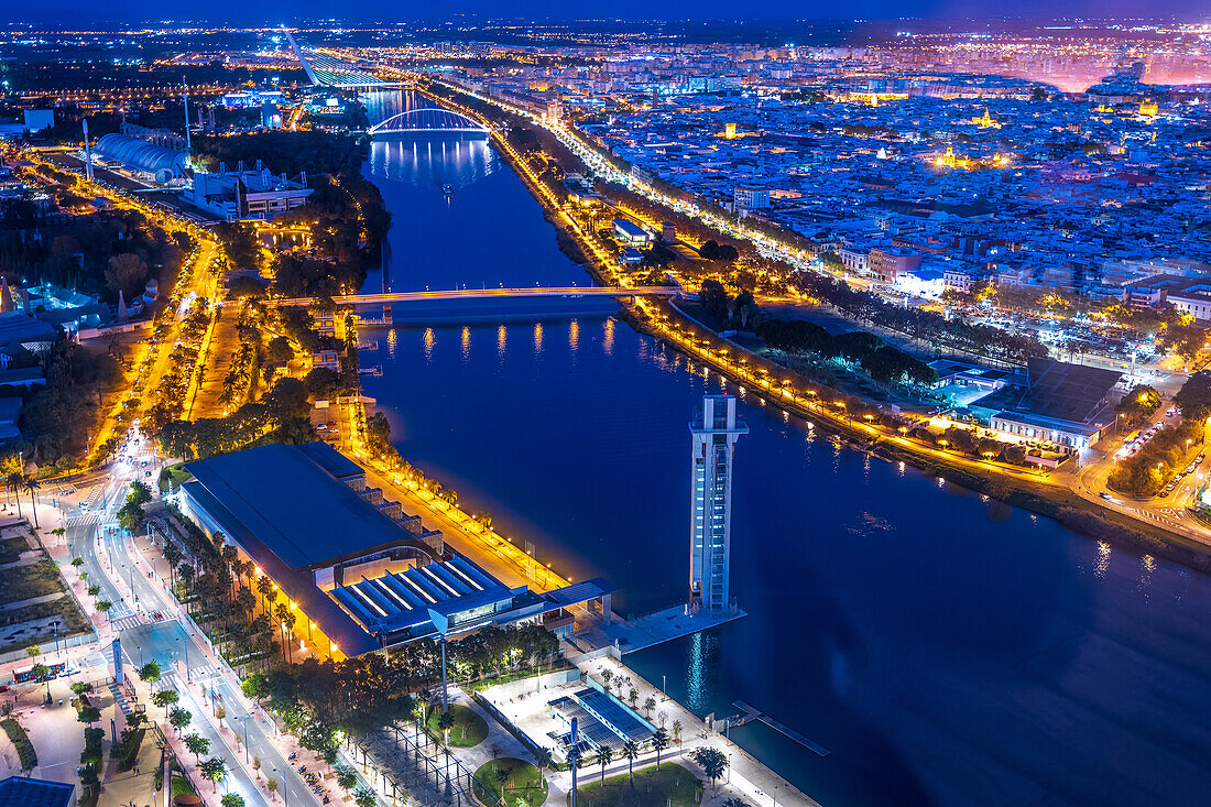 Glowing lights illuminate the Guadalquivir River in Seville, showcasing the Schildler Tower and several notable bridges at night.