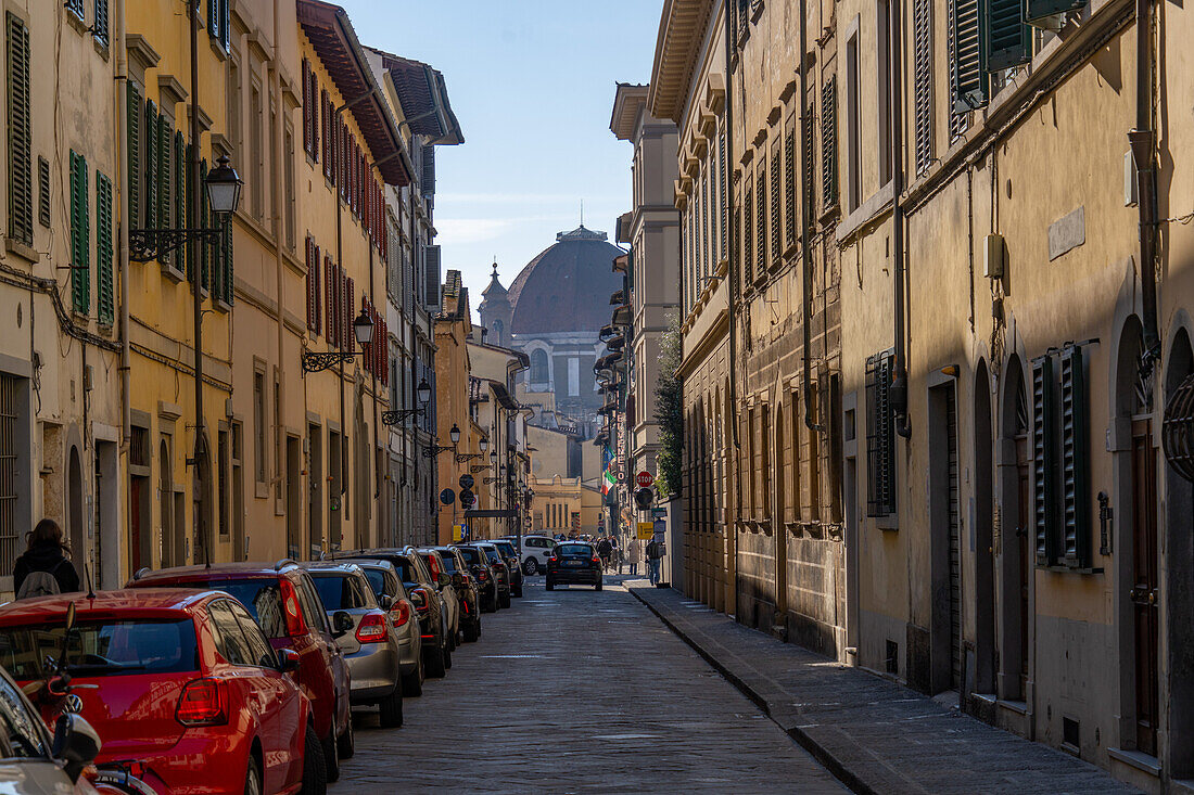 A narrow street in Florence, Italy, lined with parked cars and the dome of the Basilica of San Lorenzo behind.