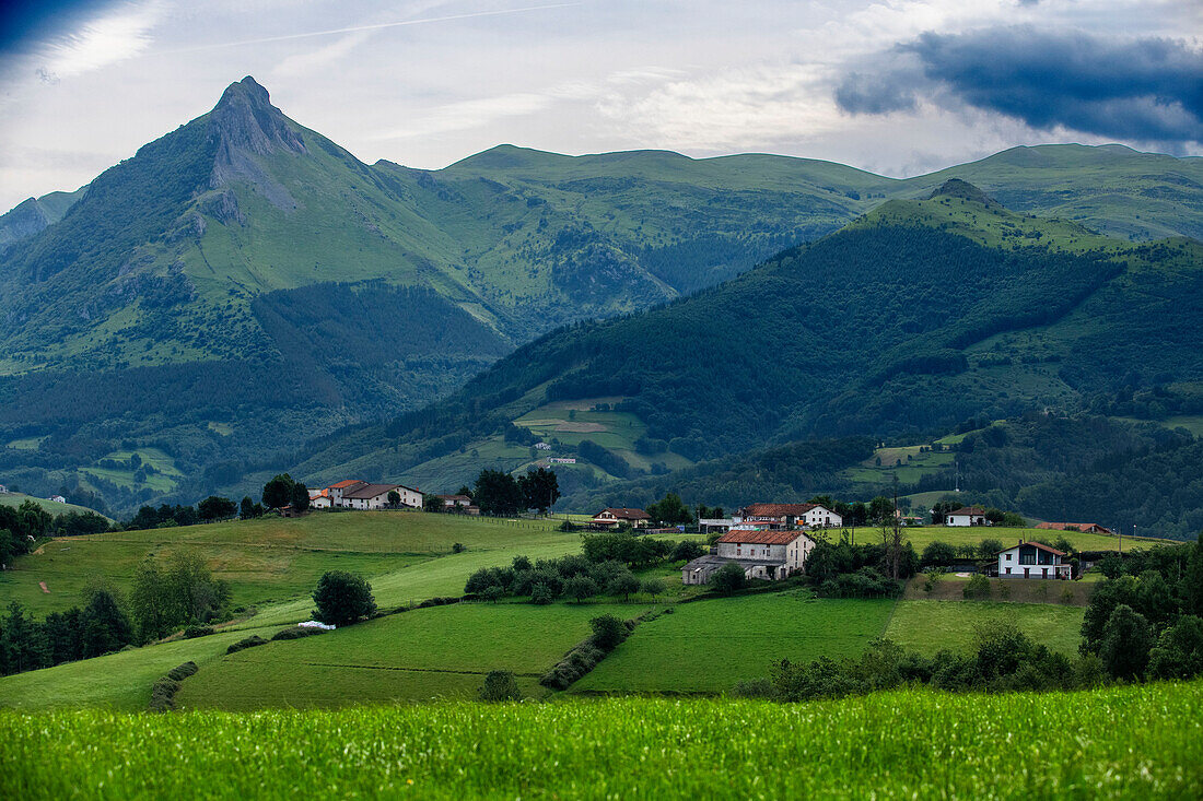 Der Berg Txindoki, grüne Wiesen, Schafherden und typische, weiß gestrichene Weiler: El Txindoki, Larrunari, steht stolz in seinem Haus in der Sierra de Aralar. Grüne Weiden, Kühe, Schafe, Pferde, Hirten. Blick von Pipas Jatetxea, Lazkaomendi, Lazcano, Goierri, Baskisches Hochland Baskenland, Euskadi Spanien.