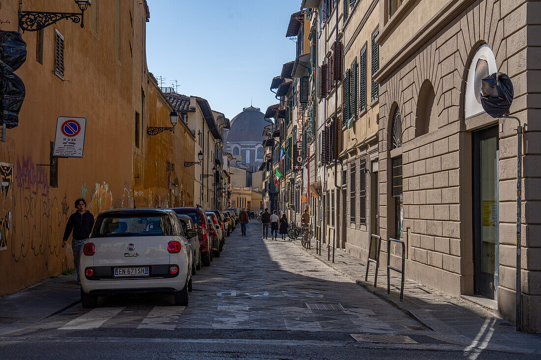 A narrow street in Florence, Italy, lined with parked cars and the dome of the Basilica of San Lorenzo behind.