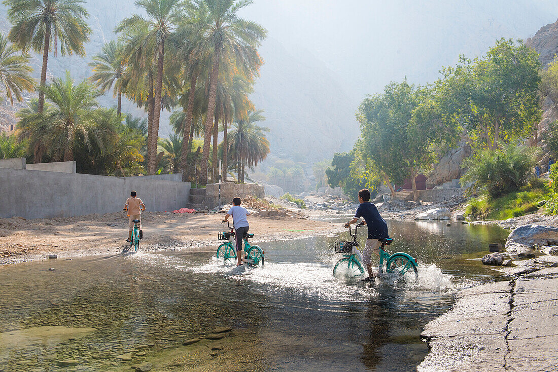 Kinder auf Fahrrädern überqueren den Fluss in der Nähe der heißen Quelle Al Thowarah (Nakhal-Quelle), Sultanat Oman, Arabische Halbinsel