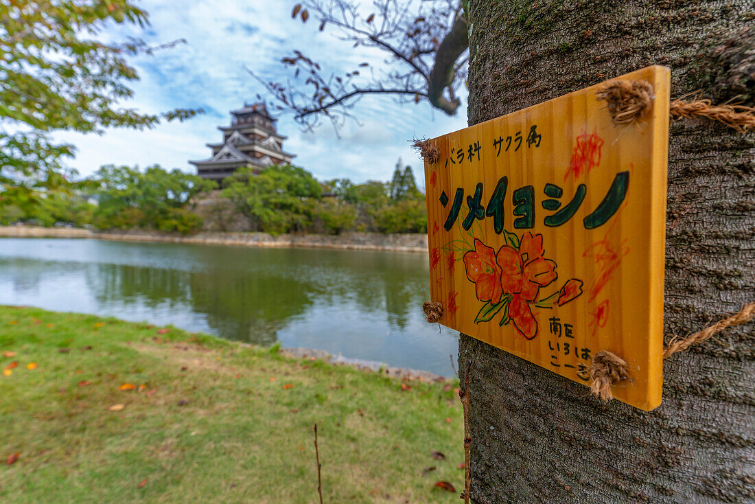 Blick auf die Burg Hiroshima mit Museum, Spiegelung im Wassergraben und Schild am Baum, Motomachi, Bezirk Naka, Hiroshima, Honshu, Japan