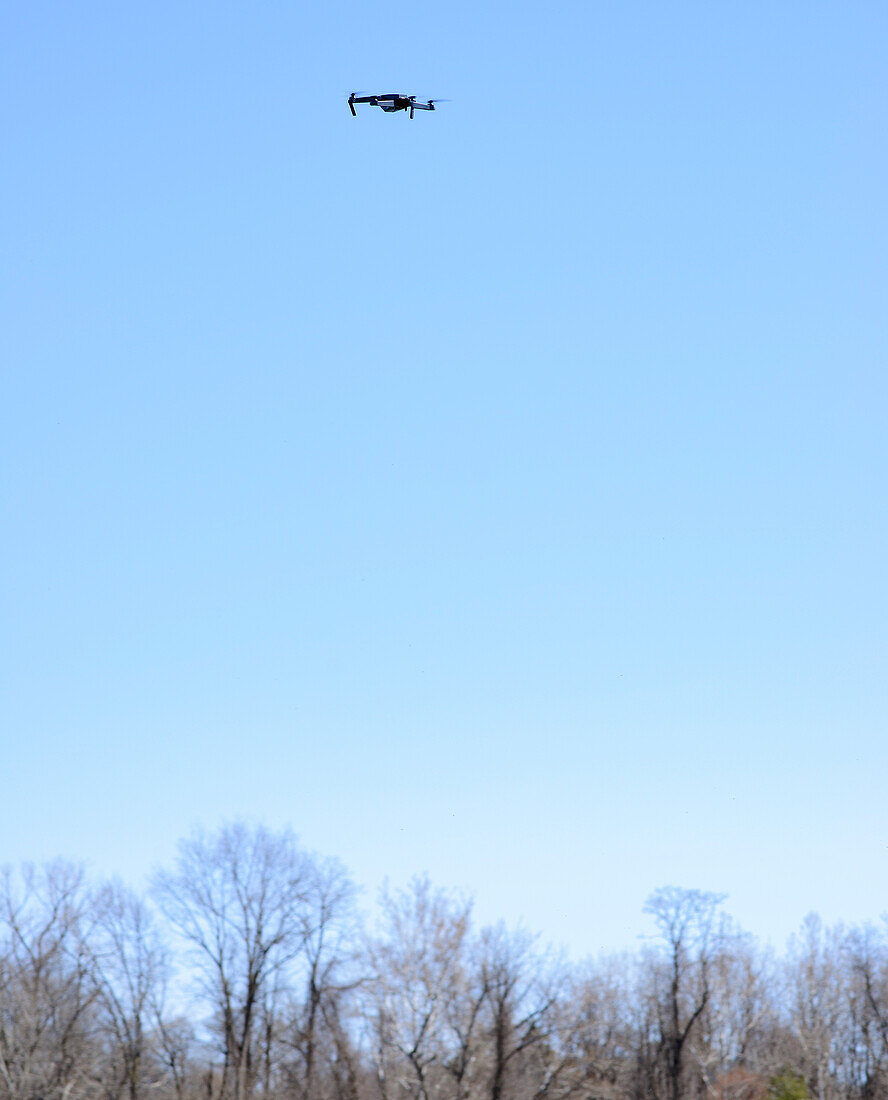 Small drone hovering in mid-air against a blue sky with bare trees on horizon