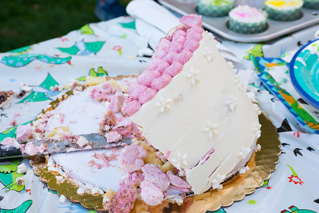 Partially-eaten vanilla birthday cake with raspberry decorations collapsing on gold foil doily with cupcakes in cupcake tin in right background