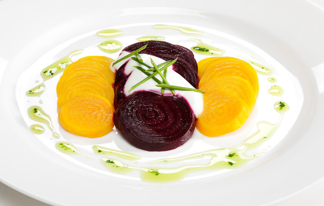 In Scheiben geschnittene gelbe und violette Rüben mit Schnittlauch und Crème fraîche, beträufelt mit Olivenöl auf weißem Teller