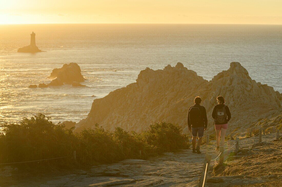 Frankreich, Finistere, Plogoff, Wanderer bei Sonnenuntergang an der Pointe du Raz, im Hintergrund der Leuchtturm der Vieille