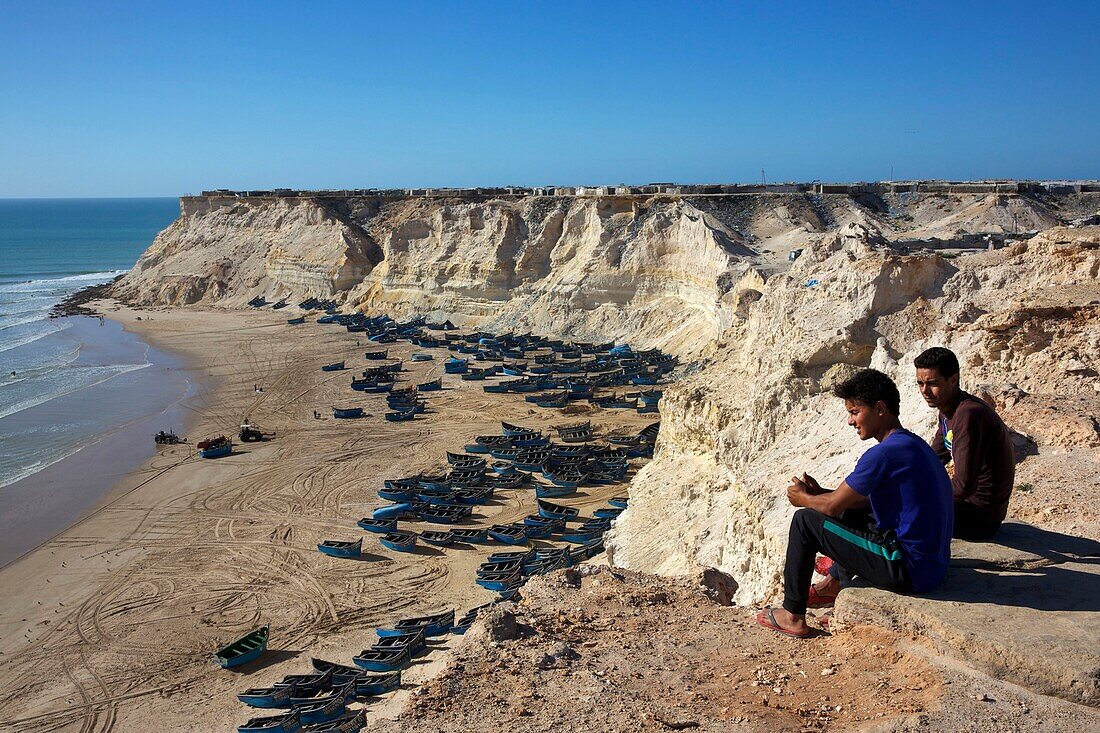 Marokko, Westsahara, Dakhla, junge Männer sitzen oberhalb der Klippe mit Blick auf den Strand von Araiche und die Fischerboote