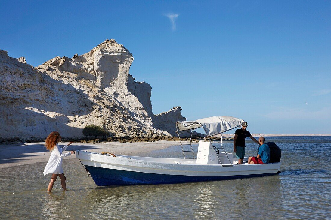 Marokko, Westsahara, Dakhla, marokkanische Frau mit Füßen im Wasser der Lagune und marokkanische Männer in einem Boot vor der Dracheninsel