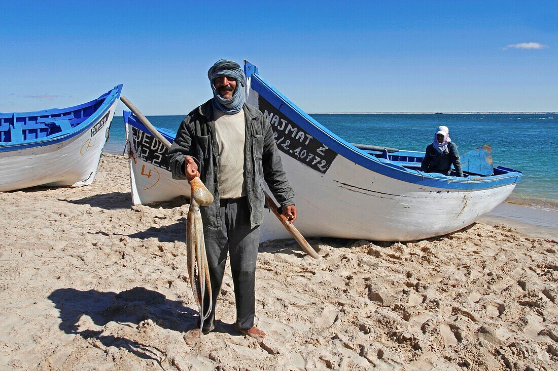 Marokko, Westsahara, Dakhla, Fischer mit einem Oktopus in der Hand vor einem Boot am Strand