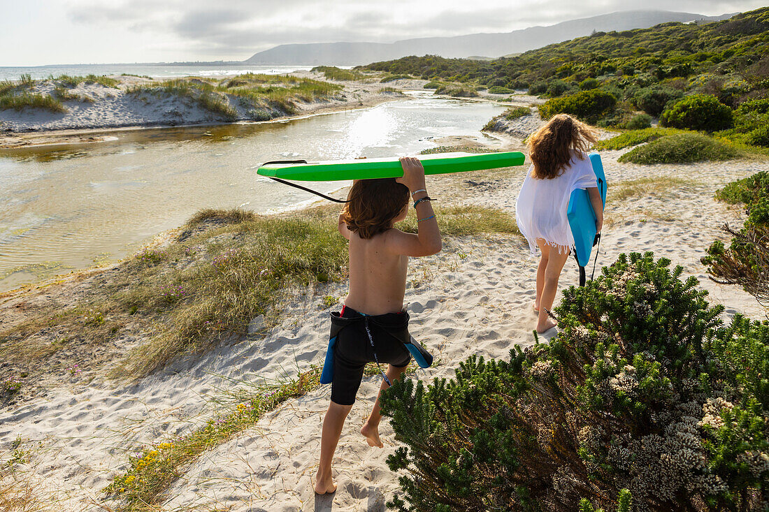 Südafrika, Hermanus, Bruder und Schwester gehen mit Bodyboards am Strand spazieren